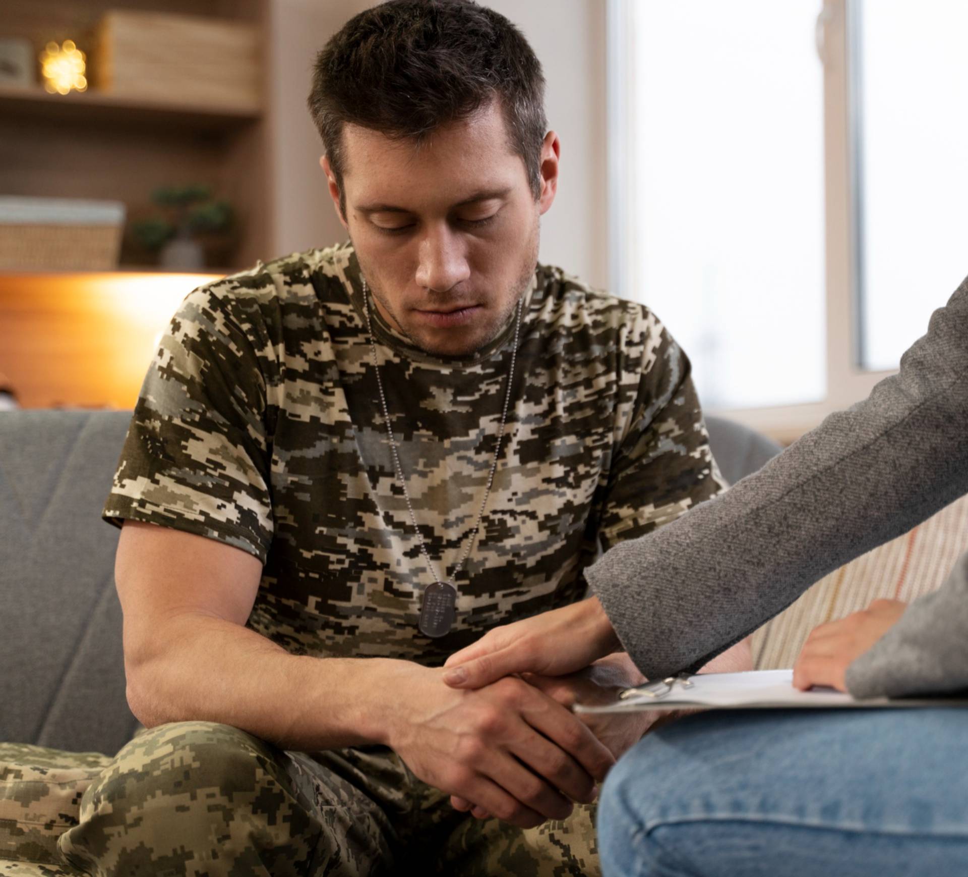 Person in camouflage sitting on couch, holding hands with individual in gray sweater.